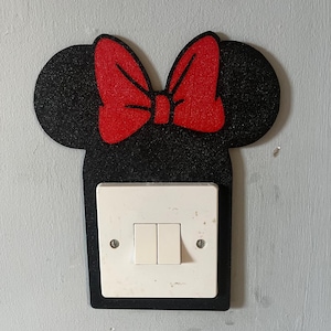 May include: A black glittery light switch cover shaped like a mouse head with large round ears and a bright red bow centred at the top. The cover surrounds a white rectangular double light switch with two vertical toggles and two screws.