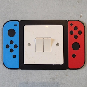 May include: A black plastic light switch cover plate featuring a blue controller-style attachment on the left side and a red controller-style attachment on the right side, surrounding a standard white double-toggle light switch.