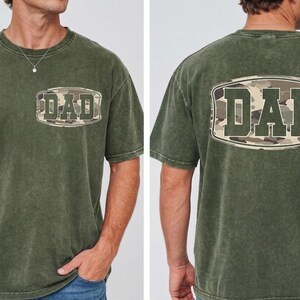 May include: Olive green t-shirt with the word "DAD" in a camouflage design on the front and back. The shirt has a crew neck and short sleeves. The fabric appears to be a soft, washed cotton.