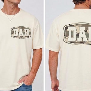 May include: Cream-colored t-shirt with the word "DAD" in a camouflage design on the front and back. The shirt has short sleeves and a crew neck. The camouflage design features shades of green, brown, and tan.
