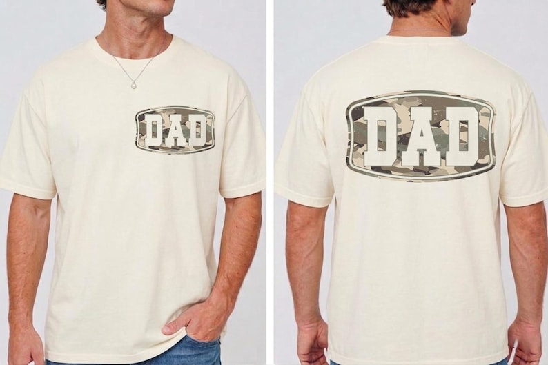 May include: Cream-colored t-shirt with the word "DAD" in a camouflage design on the front and back. The shirt has short sleeves and a crew neck. The camouflage design features shades of green, brown, and tan.