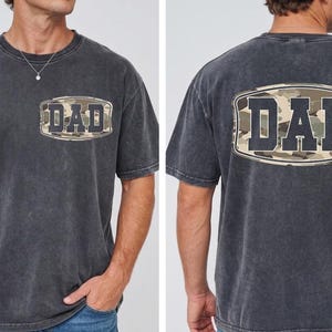 May include: Dark gray t-shirt with a camouflage design and the word "DAD" printed on the front and back. The shirt has a classic crew neck and short sleeves, perfect for casual wear.