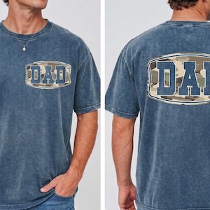 May include: A blue short-sleeved t-shirt with the word "DAD" in a camouflage design on the front and back. The shirt has a vintage, washed look.