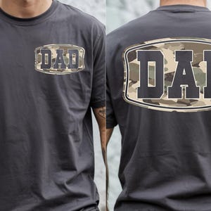 May include: Charcoal gray t-shirt with the word "DAD" in a camouflage design on the front and back. The text is inside a white and black oval. The shirt is short-sleeved and made of a soft material.