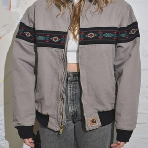 Puede incluir: Chaqueta bomber gris con una banda negra y estampada en el pecho y las mangas. La chaqueta tiene cierre de cremallera y pu&ntilde;os y dobladillo negros. Un peque&ntilde;o parche con el logo es visible en la parte delantera.