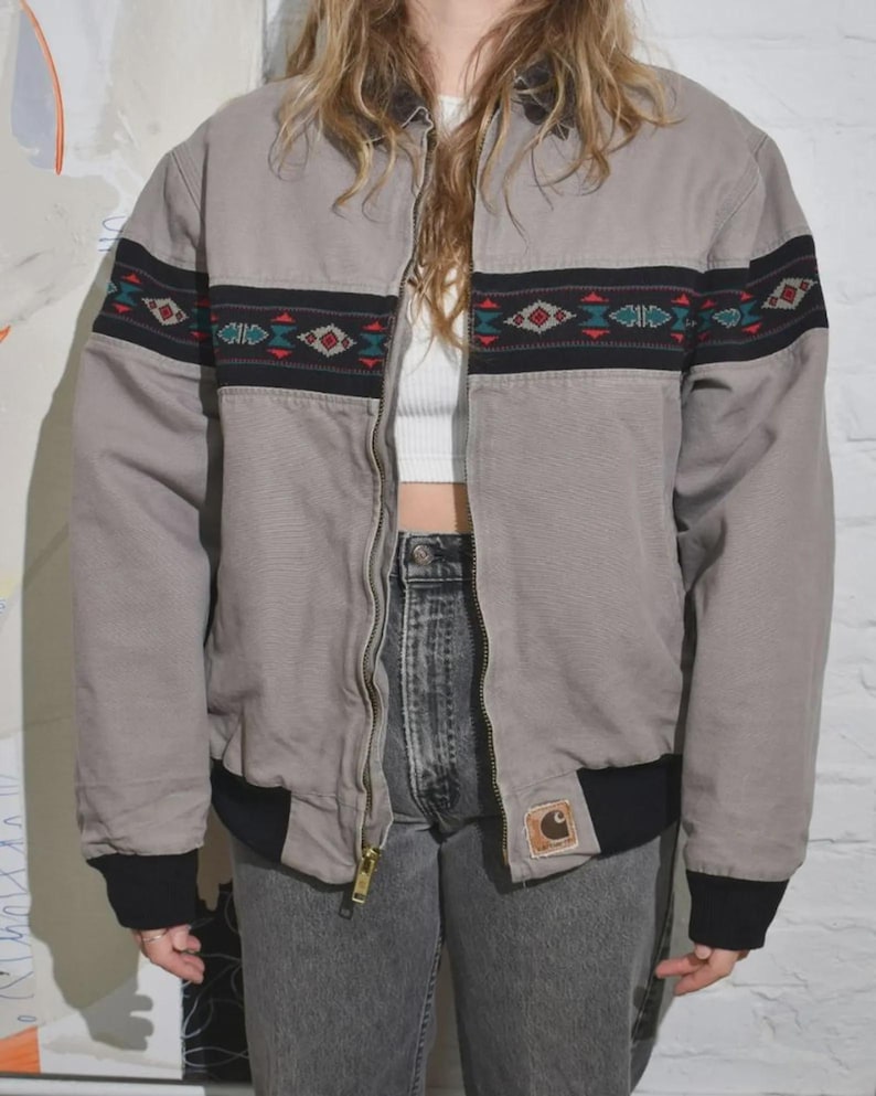 Puede incluir: Chaqueta bomber gris con una banda negra y estampada en el pecho y las mangas. La chaqueta tiene cierre de cremallera y pu&ntilde;os y dobladillo negros. Un peque&ntilde;o parche con el logo es visible en la parte delantera.
