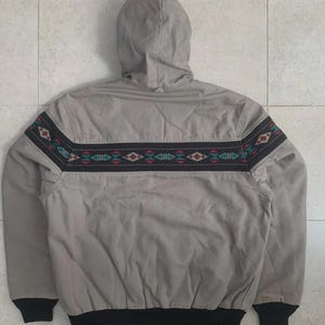 Puede incluir: Chaqueta con capucha gris claro con una banda decorativa con dise&ntilde;os geom&eacute;tricos en rojo, verde y blanco. La chaqueta tiene un dobladillo y pu&ntilde;os de canal&eacute; negros.