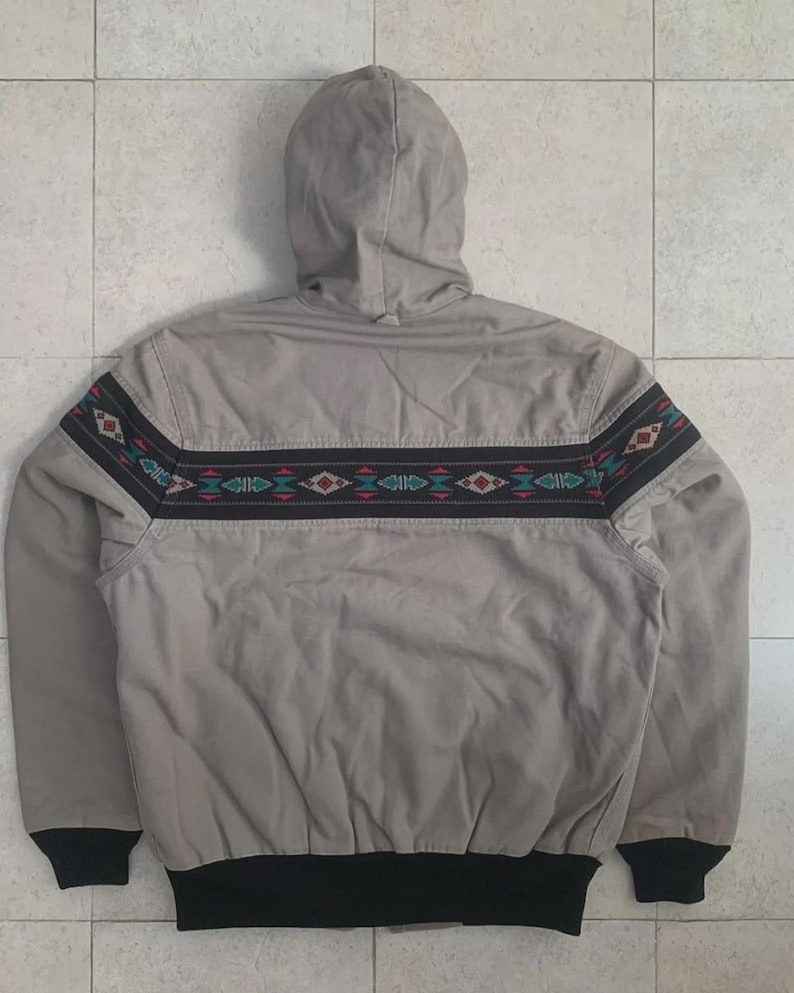 Puede incluir: Chaqueta con capucha gris claro con una banda decorativa con dise&ntilde;os geom&eacute;tricos en rojo, verde y blanco. La chaqueta tiene un dobladillo y pu&ntilde;os de canal&eacute; negros.