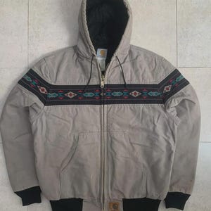 Puede incluir: Chaqueta con capucha gris claro con un dise&ntilde;o negro y estampado en el pecho. La chaqueta tiene una cremallera completa, dos bolsillos delanteros y pu&ntilde;os y dobladillo negros. La capucha est&aacute; forrada con tela negra.