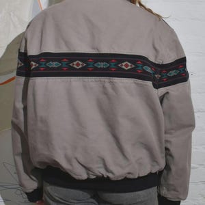 Puede incluir: Chaqueta bomber beige con una banda decorativa en la espalda. La banda presenta dise&ntilde;os geom&eacute;tricos rojos, turquesa y blancos sobre un fondo negro. La chaqueta tiene cuello marr&oacute;n y pu&ntilde;os y dobladillo negros.