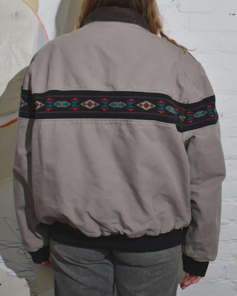 Puede incluir: Chaqueta bomber beige con una banda decorativa en la espalda. La banda presenta dise&ntilde;os geom&eacute;tricos rojos, turquesa y blancos sobre un fondo negro. La chaqueta tiene cuello marr&oacute;n y pu&ntilde;os y dobladillo negros.