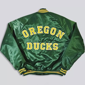 May include: Green satin jacket with the words "OREGON DUCKS" in yellow block letters. The jacket has yellow and green striped trim on the collar, cuffs, and waist. The jacket is a classic varsity style.