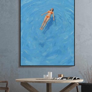 May include: An oil painting depicts a person floating in a bright blue pool. The person is wearing an orange bikini. The painting is framed in a black frame and hangs on a gray wall. The painting is above a wooden table.