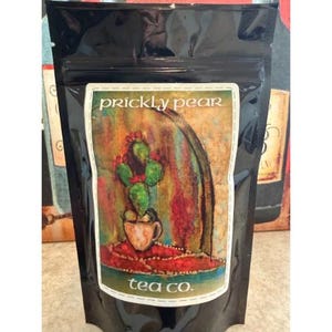 May include: Black bag of Prickly Pear Tea Co. tea. The label features a colorful illustration of a cactus with a cup, set against a backdrop of red, orange, and green hues. The words "prickly pear" and "tea co." are printed on the label.