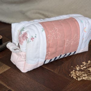 May include: A rectangular, quilted pencil case with a white zipper. The case features panels of white fabric with floral and bunny prints, and a central peach-colored panel with bunny illustrations. The bottom is pink, and the case is on a wooden surface.