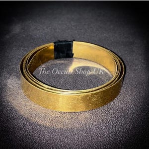 Puede incluir: Una tira de metal enrollada de color dorado, posiblemente latón u oro, con una banda negra que asegura un extremo. The Occult Shop UK está impreso en la superficie. La tira de metal está sobre una superficie oscura y texturizada.