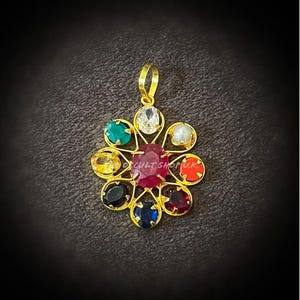 May include: A gold-colored pendant with a flower-like design. The center features a large, oval ruby, surrounded by smaller gemstones in various colors, including green, yellow, blue, and red. The pendant has a loop at the top for a chain.