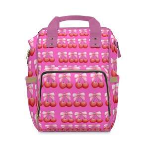 Puede incluir: Mochila rosa con un patrón repetido de cerezas atadas con lazos blancos. La bolsa tiene un asa superior, un bolsillo frontal y bolsillos laterales. El asa y el ribete son de un rosa más oscuro.
