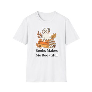 Puede incluir: Una camiseta blanca de manga corta con un gráfico de un fantasma con gafas en forma de corazón y una flor, sentado sobre una pila de tres libros naranjas rodeados de hojas de otoño y bellotas, con el texto "Books Makes Me Boo-tiful".