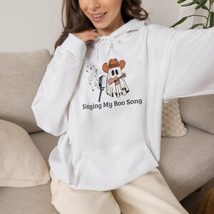 Puede incluir: Una sudadera con capucha blanca que presenta un gráfico de un fantasma con sombrero de vaquero marrón, tocando una guitarra acústica y junto a un micrófono. Notas musicales flotan cerca y el texto Singing My Boo Song está impreso.