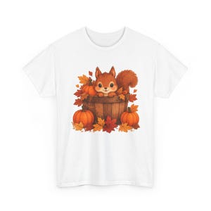 Puede incluir: Una camiseta blanca con un estampado gráfico de una ardilla marrón sentada dentro de un barril de madera. La ardilla está rodeada de calabazas naranjas y hojas de otoño coloridas en tonos rojos, amarillos y naranjas, creando una escena.