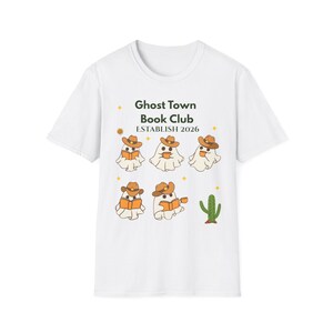 Puede incluir: Una camiseta blanca con el texto Ghost Town Book Club ESTABLISH 2026 en verde oscuro y negro. El gráfico incluye cinco fantasmas de dibujos animados con sombreros de vaquero marrones, leyendo libros o sosteniendo tazas, y un cactus verde.