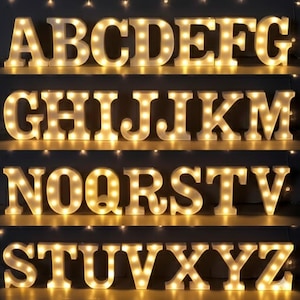 Puede incluir: Letras de marquesina blancas e iluminadas del alfabeto, dispuestas en filas. Cada letra está iluminada con pequeñas bombillas de tonos cálidos. Las letras se muestran en estantes, creando una exhibición decorativa y llamativa.