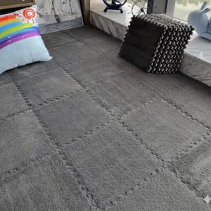 May include: Gray interlocking foam floor mats. The mats are arranged to cover a floor area. A pillow with a rainbow and sun design is in the upper left corner. A stack of black and white mats is in the upper right corner.