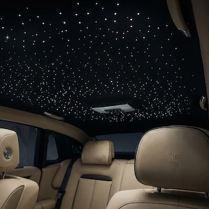 Op de afbeelding: Interieur van een luxe voertuig met een zwart hemelbekleding met ingebedde vezeloptische lichten die een sterrenhemel simuleren. Zandkleurige leren stoelen en hoofdsteunen zijn zichtbaar, met het Rolls-Royce-logo op de hoofdsteun.