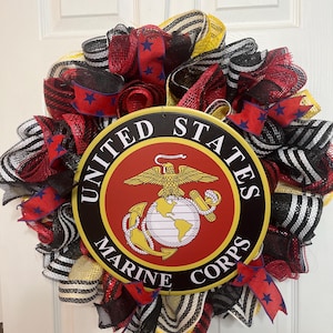 May include: A decorative wreath with a central emblem of the United States Marine Corps. The wreath is made of red, black, white, and yellow ribbons, with blue star-patterned ribbon accents. The emblem is a circular design with the words "United States Marine Corps".
