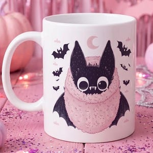 Peut inclure: Un mug en céramique blanche orné d'une illustration graphique représentant une chauve-souris noire duveteuse aux grands yeux et aux crocs pointus, enveloppée dans un col texturé rose pâle. Le motif inclut un croissant de lune et des chauves-souris.