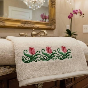 Könnte beinhalten: Cremefarbenes Badetuch mit einem gestickten Blumendesign aus drei roten Tulpen und grünen Blättern. Das Handtuch liegt gefaltet auf einer Badezimmer-Arbeitsplatte, mit einem Spiegel und Kronleuchter im Hintergrund.