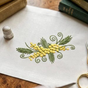 Op de afbeelding: Een witte linnen tafelloper met een geborduurd ontwerp van gele mimosa bloemen en groene bladeren. Een zilveren vingerhoed en een gouden schaar liggen op de tafel. Op de achtergrond liggen verschillende antieke boeken.