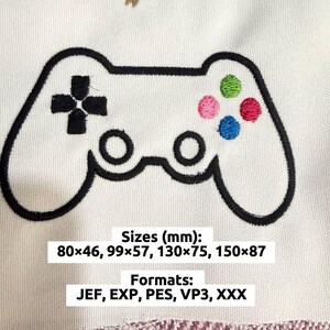 May include: White fabric with a black embroidered video game controller design. The controller has a cross-shaped directional pad, two circular buttons, and four colored buttons: green, pink, blue, and red. The text "Sizes (mm): 80x46, 99x57, 130x75, 150x87" and "Formats: JEF, EXP, PES, VP3, XXX" are also visible.