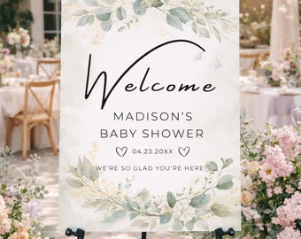 May include: A welcome sign for Madison's baby shower, displayed on a black easel. The sign features elegant script and floral accents in green and white. The date is 04.23.20XX, with the text "We're so glad you're here!"