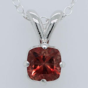 May include: A silver pendant necklace featuring a square-cut, reddish-orange gemstone. The gemstone is set in a four-prong setting and hangs from a delicate silver chain. The pendant has a simple, elegant design.