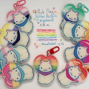 Può includere: Una collezione di portachiavi in acrilico glitterato con bandiera Pride. Ogni portachiavi presenta un design di gatto bianco all'interno di una forma a cuore, con vari schemi di colori arcobaleno. I portachiavi misurano circa 7 cm e hanno un fermaglio rosso.