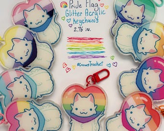 May include: A collection of Pride Flag glitter acrylic keychains. Each keychain features a white cat design within a heart shape, with various rainbow flag color schemes. The keychains are approximately 2.76 inches in size and have a red clasp.