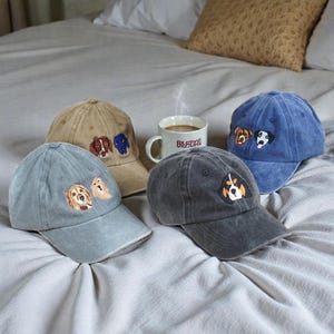 Peut inclure: Quatre casquettes de baseball de couleurs variées, dont bleu clair, beige et gris foncé. Chaque casquette est ornée d'une broderie de chien. Une tasse blanche avec le mot "Bestie" est également visible.