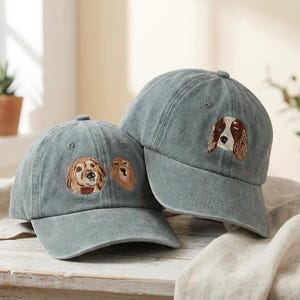 May include: Two blue-green baseball caps with embroidered dog faces. One cap features a golden retriever and a second dog, while the other displays a spaniel. The caps are on a wooden surface.