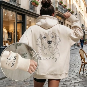 Może przedstawiać: Kremowa bluza z kapturem z rysunkiem linii przedstawiającym pysk psa i imieniem "Bailey". Lewy mankiet rękawa przedstawia grafikę psa i serca z napisem "DOG MOM". Bluza z kapturem jest noszona przez osobę idącą po brukowanej ulicy.
