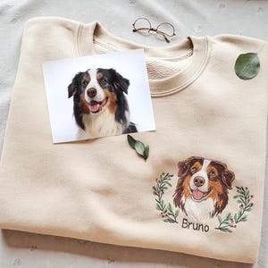 Op de afbeelding: Beige crewneck sweatshirt met een geborduurd hondenportret en de naam "Bruno" eronder. Een foto van de hond is op de sweatshirt geplaatst. De hond heeft bruin, zwart en wit haar. Een bril en een blad liggen op de sweatshirt.