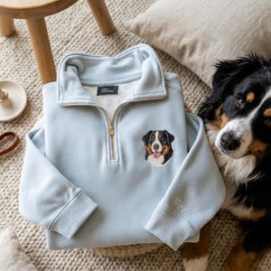 May include: A pale blue quarter-zip sweatshirt with a gold zip and an embroidered Bernese Mountain Dog on the chest. The sleeve is embroidered with the name "Finn". The sweatshirt is displayed with a dog.