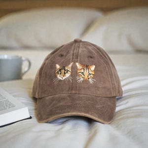 Peut inclure: Casquette de baseball marron avec des visages de chats brodés. La casquette a une visière incurvée et une fermeture à sangle en tissu. Les visages de chats sont détaillés avec du fil marron, beige et blanc.