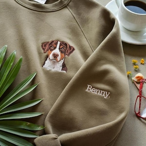 Può includere: Felpa verde oliva con un ritratto di cane ricamato sul petto. Il cane ha segni marroni, bianchi e neri. Il nome "Benny" è ricamato sulla manica. Una tazza di caffè e occhiali sono sullo sfondo.
