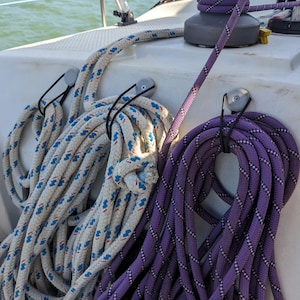 May include: Close-up of coiled boat ropes in white with blue accents and a purple rope. The ropes are hanging on a white boat surface. A boat winch is visible in the background. The scene suggests nautical or boating themes.