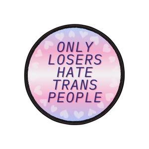 May include: Round patch with a black border. The patch features a gradient background of pink and light blue with white hearts. The text "ONLY LOSERS HATE TRANS PEOPLE" is in dark blue.