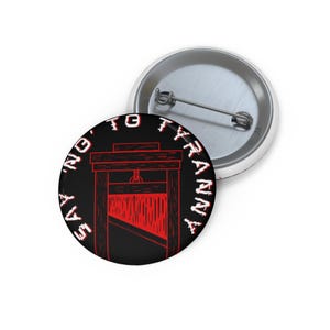Könnte beinhalten: Ein runder Button mit schwarzem Hintergrund und einer roten Guillotine-Illustration. Der Text "SAY NO TO TYRANNY" ist in Weiß am Rand des Buttons geschrieben. Ein zweiter Button ist im Hintergrund sichtbar.