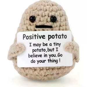 Op de afbeelding: Een gehaakt aardappelvormig pluche speelgoed in beige, met zwarte knoopogen en een gestikte mond. Het houdt een wit bord vast met de tekst "Positive potato" en een motiverende boodschap.