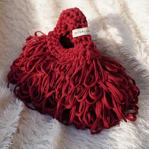 May include: A handmade, red crocheted bag with a unique design. The bag features a looped fringe and a label that reads "SINALUN". The bag is made of a soft, textured material and is displayed on a fluffy white surface.